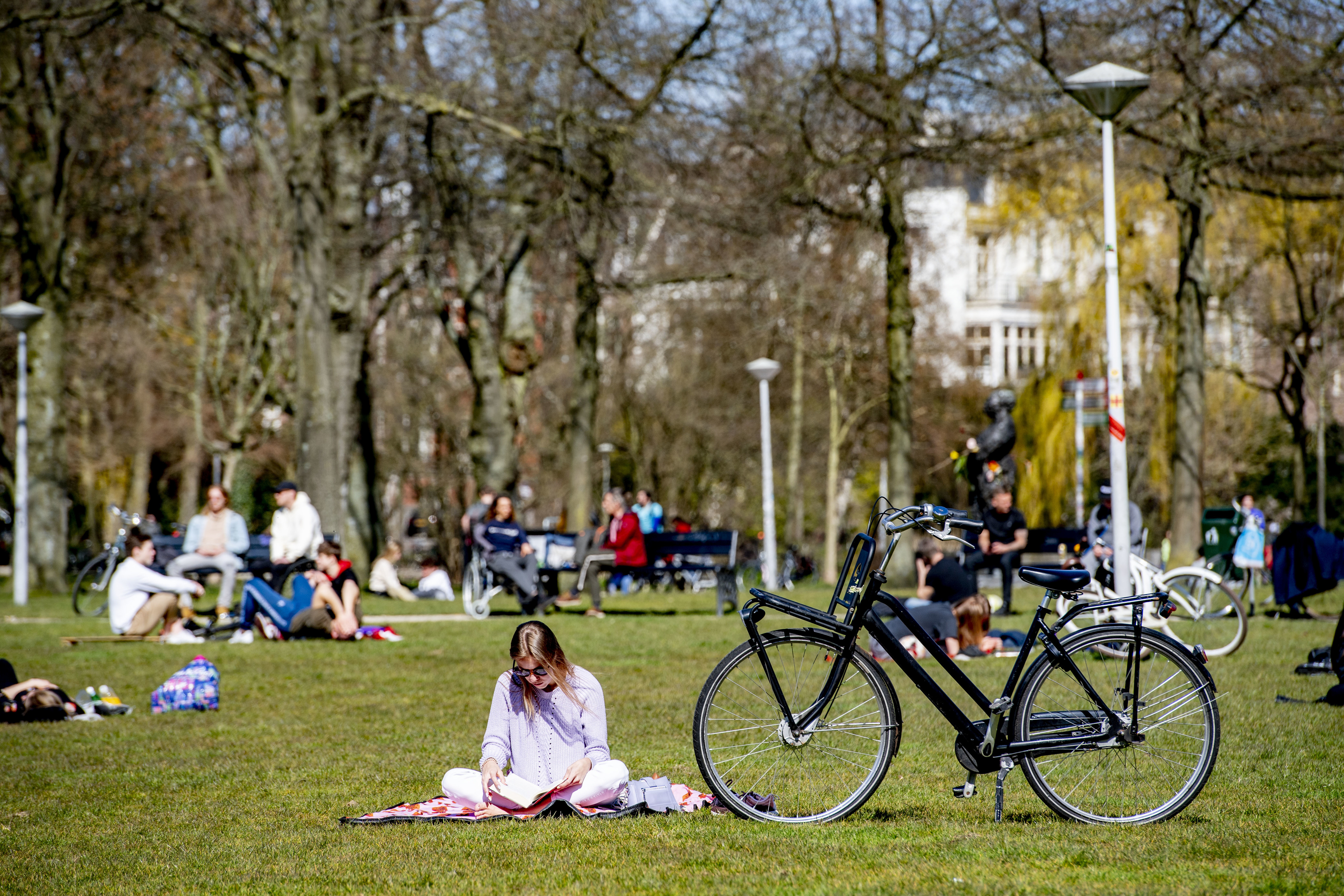 Vondelpark A woman sits cross legged and reads a book in the sunshine during a spring day at the Vondelpark in Amsterdam. She is sat next to her black bicycle with trees and other park users in the background.